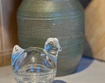 Könnte beinhalten: Ein durchsichtiger Glasbehälter in Form eines Huhns mit einem strukturierten, korbähnlichen Boden. Der Behälter steht auf einer weißen Arbeitsplatte neben einer grünen Keramikvase.