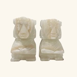 May include: A pair of white alabaster bookends featuring carved faces of Mayan figures. The bookends are detailed with intricate carvings and have a smooth, polished finish.