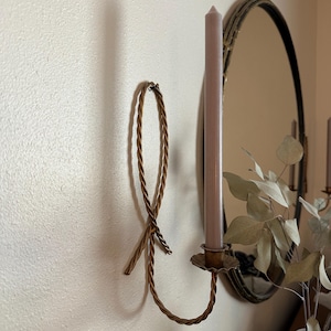 K&ouml;nnte beinhalten: Ein goldfarbener Kerzenhalter aus Metall mit einem Seil-Design. Der Kerzenhalter ist an einer Wand befestigt und hat eine einzelne Kerze darin. Die Kerze ist helllila.