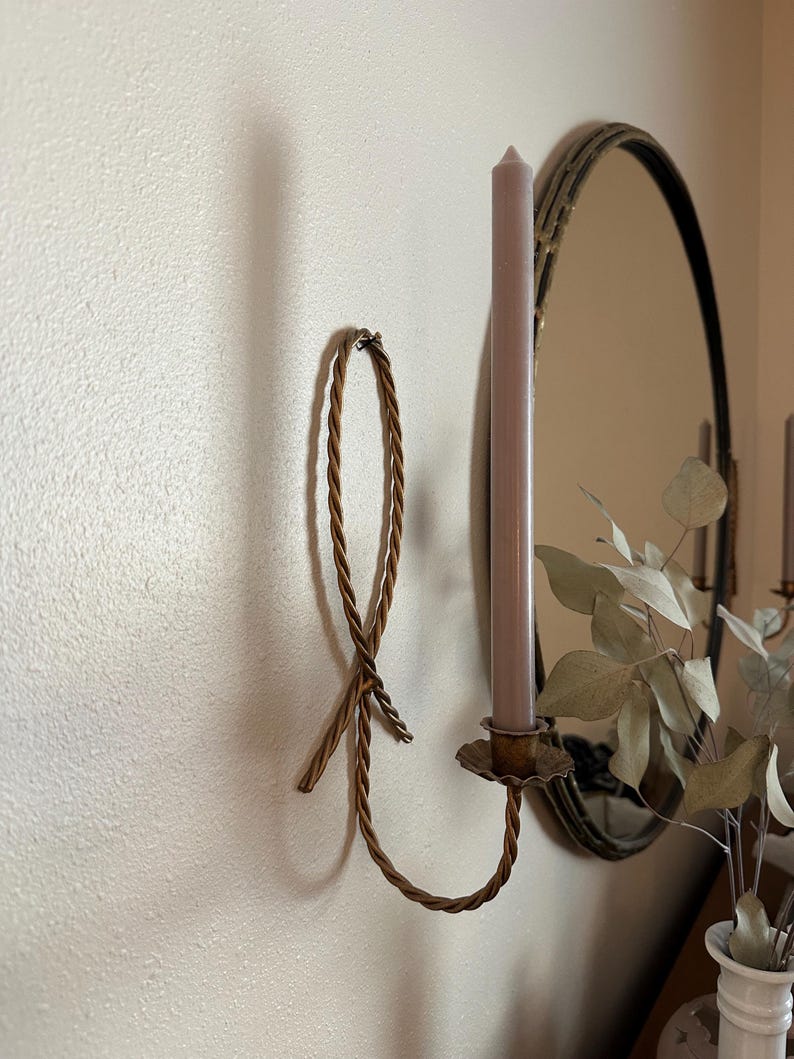 K&ouml;nnte beinhalten: Ein goldfarbener Kerzenhalter aus Metall mit einem Seil-Design. Der Kerzenhalter ist an einer Wand befestigt und hat eine einzelne Kerze darin. Die Kerze ist helllila.