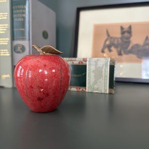 Könnte beinhalten: Ein roter Marmorapfel mit einem goldenen Blattstiel. Der Apfel liegt auf einer grauen Oberfläche.