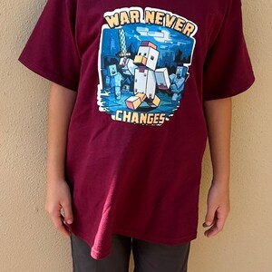 May include: A maroon short-sleeved t-shirt with a graphic print of a Minecraft character holding a sword. The text "WAR NEVER CHANGES" is printed on the shirt.