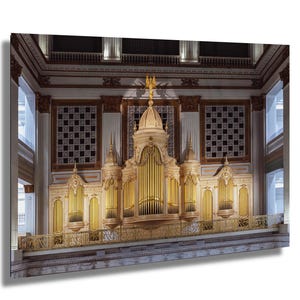 May include: A large, ornate pipe organ with gold-colored pipes and decorative elements. The organ is set against a backdrop of a high ceiling with architectural details, including windows and decorative panels. The overall color scheme is cream and gold.