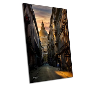 May include: A vertical print depicting a European city street. The image shows a narrow street flanked by buildings, leading to a large domed church. The sky is a mix of orange and yellow, suggesting sunrise or sunset. The street is paved with bricks.