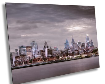 May include: A panoramic view of the Philadelphia skyline at dusk. The city's iconic skyscrapers are illuminated against a backdrop of a cloudy sky. The Delaware River is visible in the foreground.