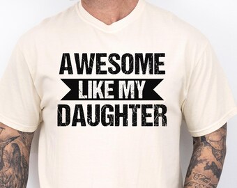 May include: Ivory-colored t-shirt with the text "AWESOME LIKE MY DAUGHTER" in black. The shirt has a crew neck and short sleeves. The word "IVORY" is printed at the bottom right.