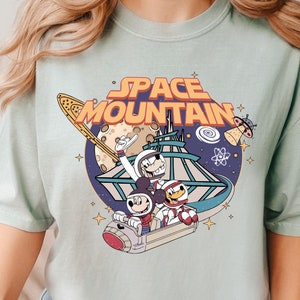 May include: A light blue t-shirt with a retro-style graphic of Mickey Mouse and friends riding a roller coaster in space. The graphic includes the text "SPACE MOUNTAIN" in orange and yellow letters.