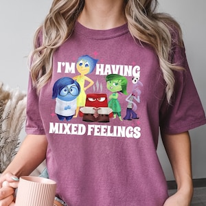 May include: Berry-colored t-shirt featuring the phrase "I'M HAVING MIXED FEELINGS" with cartoon characters. The design includes characters representing emotions like joy, sadness, anger, disgust, and fear. The shirt is a comfortable, casual wear.