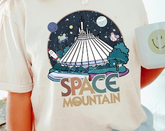 May include: A white t-shirt with a retro-style graphic of Space Mountain, a roller coaster ride at Disney World. The graphic features a colorful illustration of the ride, with stars, planets, and a rocket ship. The text "SPACE MOUNTAIN" is written in a colorful, retro font.