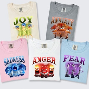 May include: Five t-shirts featuring characters from the movie Inside Out. The shirts are white, light blue, pink, cream, and gray. The shirts have the words "Joy", "Sadness", "Anger", "Fear", and "Anxiety" printed on them. Each shirt features a different character from the movie.