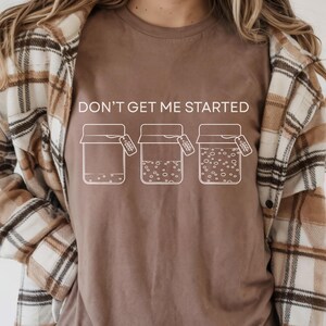 May include: Espresso-colored t-shirt with the phrase "DON'T GET ME STARTED" above three jars with tags. The jars are in different stages of filling. The person is wearing a plaid shirt.