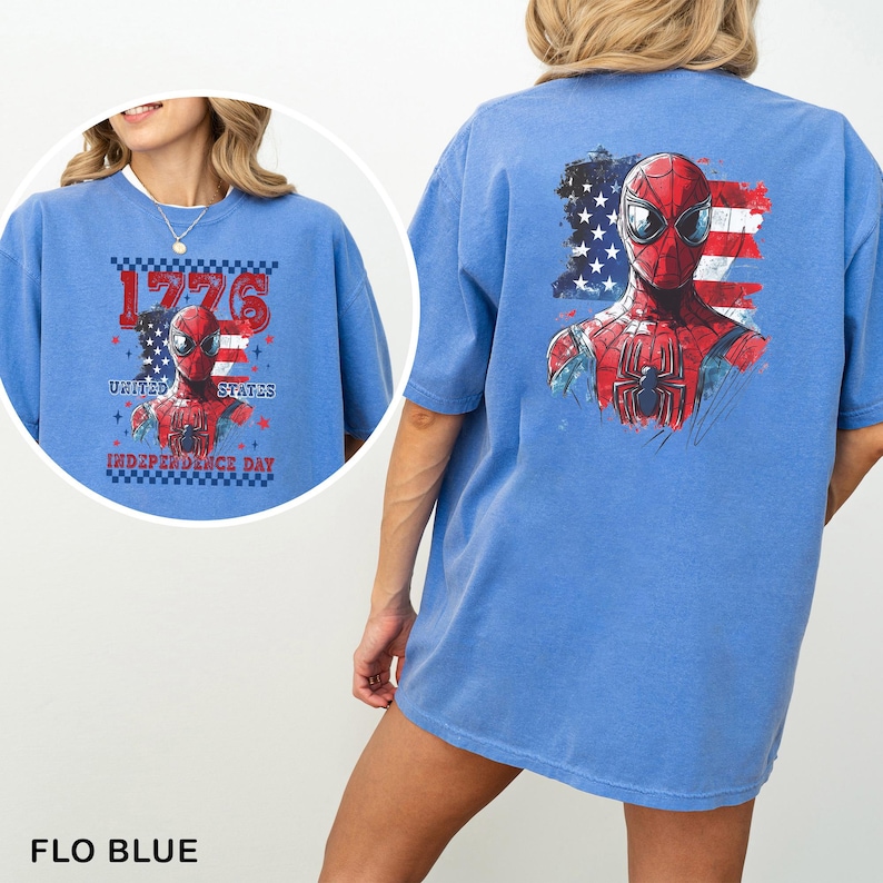 May include: A blue t-shirt featuring a graphic of a superhero mask and chest emblem overlaid on a distressed American flag. The design includes the text 1776 UNITED STATES INDEPENDENCE DAY. The back of the shirt displays the same superhero graphic.