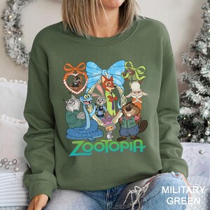 Op de afbeelding: Militairgroen sweatshirt met een kleurrijke afbeelding van personages uit de animatiefilm "Zootopia". Het ontwerp bevat een vos, konijn en andere dieren in een decoratief kader, met het woord "Zootopia" in turquoise eronder.