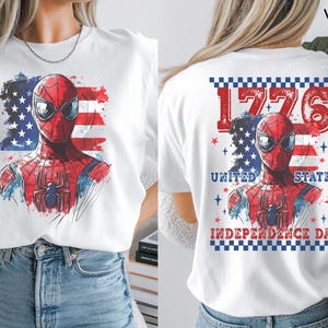 May include: A white t-shirt featuring a graphic of a red and blue superhero mask over a stylized American flag. The back design includes the text 1776, UNITED STATES, and INDEPENDENCE DAY, framed by a blue and white checkered border and red stars.