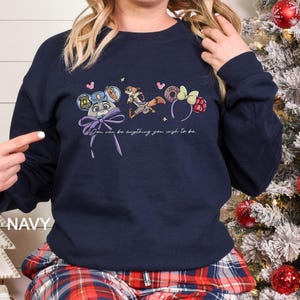 May include: Navy blue sweatshirt featuring cartoon characters and the text "You can be anything you wish to be." The design includes a bunny, fox, and themed headbands. A purple bow is at the bottom of the design.