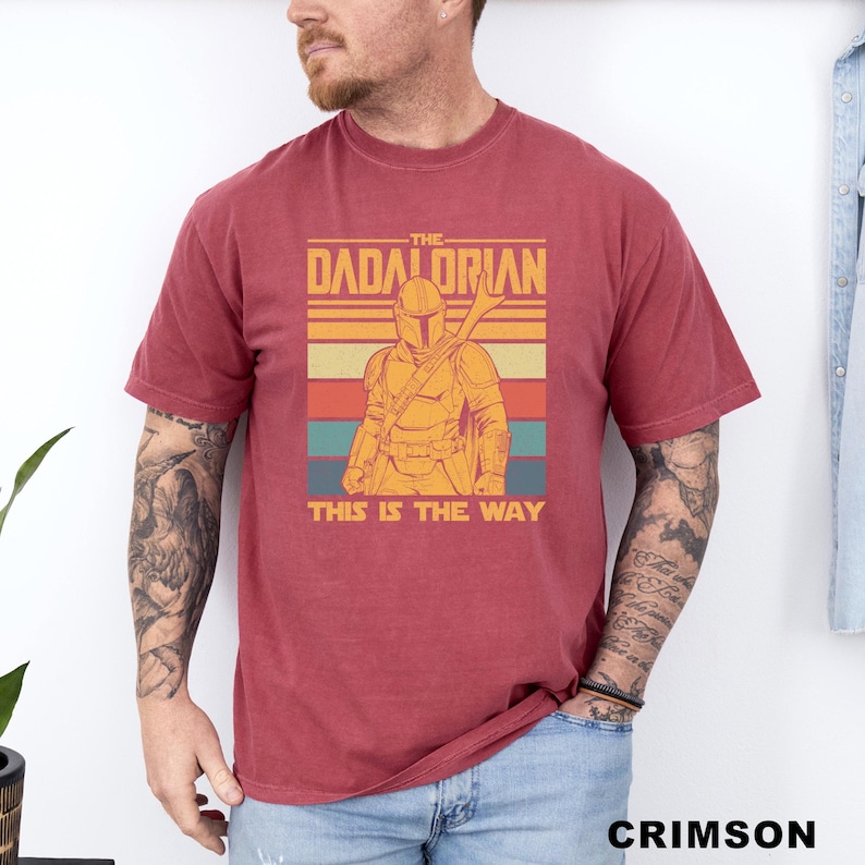 May include: Crimson-colored t-shirt featuring a retro design with the text "THE DADALORIAN" above an illustration of a Mandalorian-style character. Below the image, the text reads "THIS IS THE WAY".