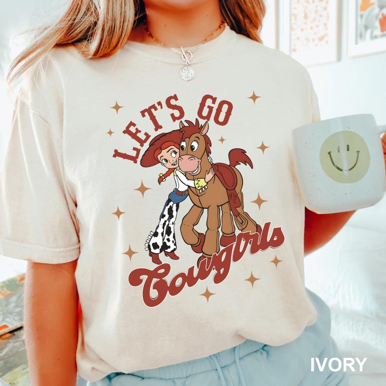 May include: A white t-shirt featuring a cartoon cowgirl and a horse graphic. The graphic reads "Let's Go Cowgirls".