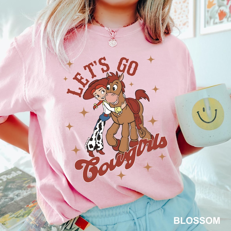 May include: Pink t-shirt featuring a cartoon cowgirl and a horse. The text "Let's Go Cowgirls" is printed on the shirt.