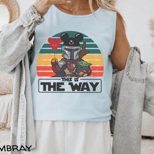 May include: Light blue tank top featuring a graphic of The Mandalorian with a Mickey Mouse head, holding a red heart balloon, and Grogu. The design is set against a retro sunset with the text "THIS IS THE WAY".
