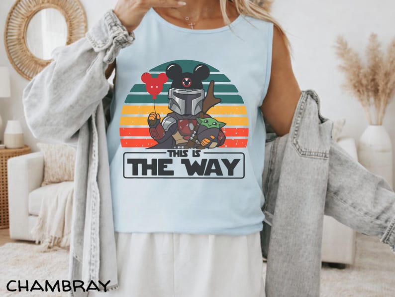 May include: Light blue tank top featuring a graphic of The Mandalorian with a Mickey Mouse head, holding a red heart balloon, and Grogu. The design is set against a retro sunset with the text "THIS IS THE WAY".