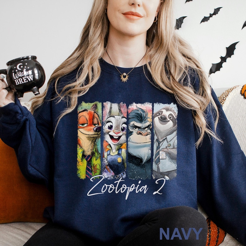 May include: Navy blue sweatshirt featuring a colorful graphic of characters from Zootopia 2. The design includes Nick Wilde, Judy Hopps, Chief Bogo, and Flash. The text "Zootopia 2" is written in white script below the characters. The word "NAVY" is printed at the bottom.