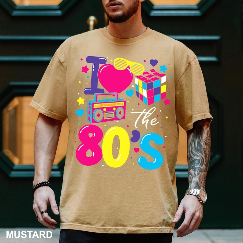 May include: Mustard-colored t-shirt with a colorful graphic that says "I love the 80's". The design includes a pink heart, a boombox, a Rubik's cube, and other 80s-themed elements.