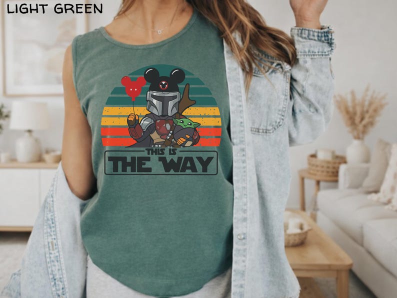 May include: Light green tank top featuring a graphic of a Mandalorian character with Mickey Mouse ears, holding a red heart balloon and Grogu. The design is set against a sunset background with the text "THIS IS THE WAY".