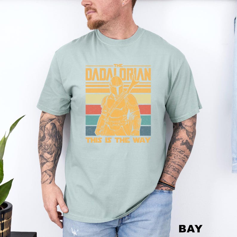 May include: A light green t-shirt featuring a retro design with the text "The Dadalorian" and "This is the Way". The design includes an illustration of a Mandalorian character in orange and yellow.