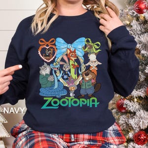 Op de afbeelding: Marineblauwe sweatshirt met een kleurrijke afbeelding van personages uit de film "Zootopia". Het ontwerp bevat een centraal frame met Judy Hopps en Nick Wilde, omringd door andere personages. Het woord "Zootopia" staat in groen en blauw onder de personages.