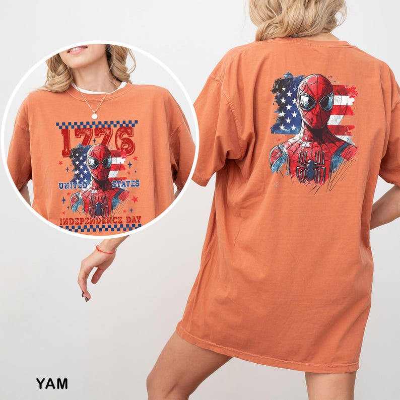May include: An orange short-sleeve t-shirt featuring a graphic of a superhero mask over a United States flag. The front design includes the text 1776 UNITED STATES INDEPENDENCE DAY. The back design shows the same superhero mask over a flag.