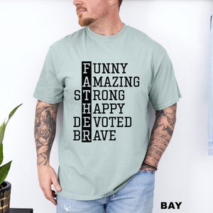 May include: A light sage green short-sleeved t-shirt featuring black text arranged in a vertical crossword style. The word FATHER is spelled vertically, with the letters forming the words FUNNY, AMAZING, STRONG, HAPPY, DEVOTED, and BRAVE.