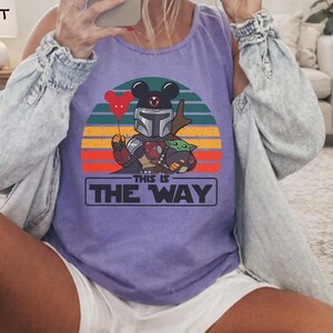 May include: Violet tank top featuring a graphic of a Mandalorian character with Mickey Mouse ears holding a heart-shaped balloon and Grogu. The design is set against a sunset backdrop with the text "THIS IS THE WAY" in a rectangular box.