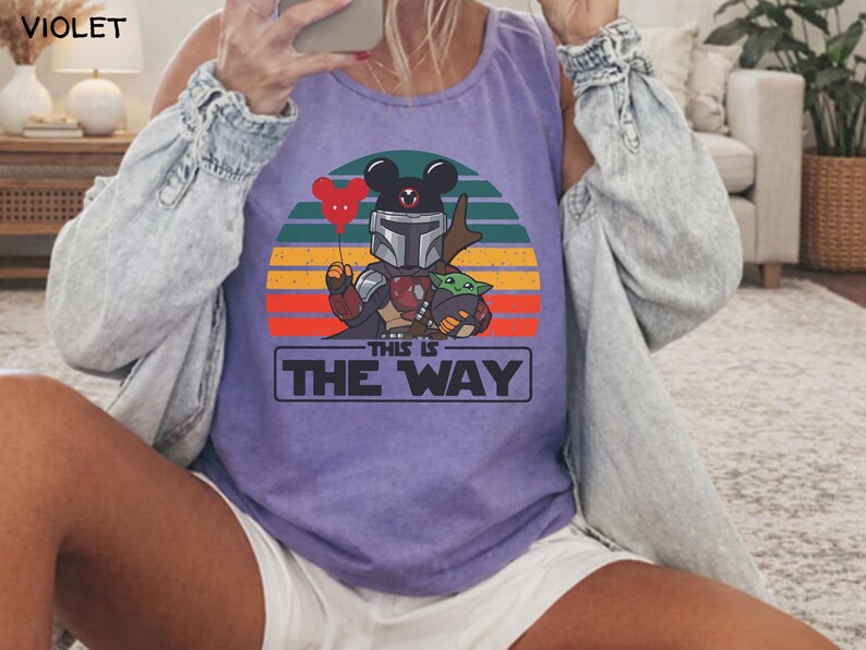 May include: Violet tank top featuring a graphic of a Mandalorian character with Mickey Mouse ears holding a heart-shaped balloon and Grogu. The design is set against a sunset backdrop with the text "THIS IS THE WAY" in a rectangular box.