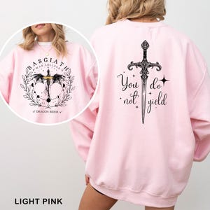 May include: Light pink sweatshirt with a black graphic on the front that says "BASGIATH WAR COLLEGE" with a dragon and sword design. The back of the sweatshirt has a sword graphic and the words "You do not yield".