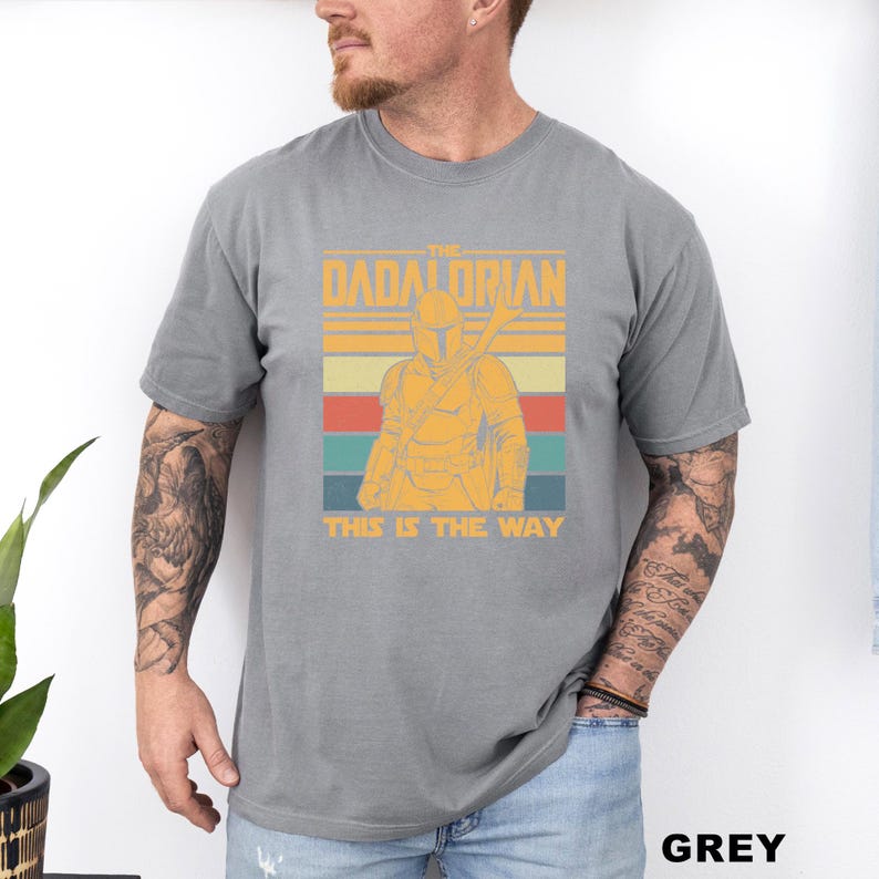 May include: Grey t-shirt featuring a retro design with the text "The Dadalorian" above an illustration of a Mandalorian character. Below the image is the phrase "This is the Way".
