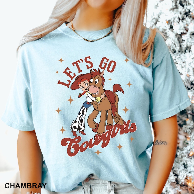 May include: Light blue t-shirt featuring a graphic of Woody and Jessie from Toy Story. The graphic says "Let's Go Cowgirls".