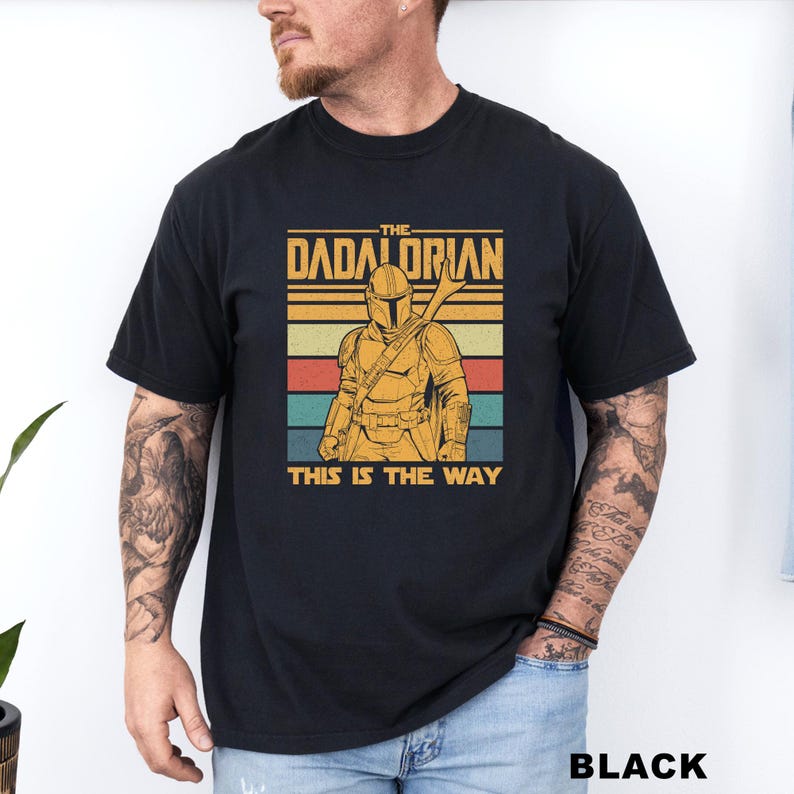 May include: Black t-shirt featuring a retro design with the text "THE DADALORIAN" above an illustration of a character in armor, with the phrase "THIS IS THE WAY" below. The design incorporates stripes of yellow, orange, red, teal, and blue.