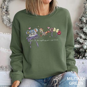 May include: An olive green sweatshirt featuring a whimsical design with cartoon characters and accessories. The design includes a mouse with a police hat, a running fox, and various headbands. The text "You can be anything you wish to be" is also included.
