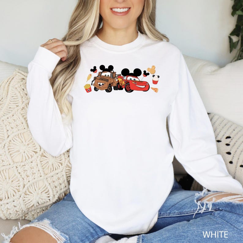 May include: White long-sleeve sweatshirt with a graphic of Mickey Mouse and two cars from the Disney movie Cars. The cars are Mater and Lightning McQueen. The graphic also includes Mickey Mouse ears, a pizza slice, french fries, and a drink.