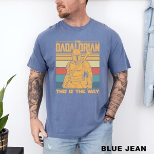 May include: Blue t-shirt featuring a graphic of a Mandalorian-style character with the text "THE DADALORIAN" and "THIS IS THE WAY" in a retro design. The shirt is a solid blue color.