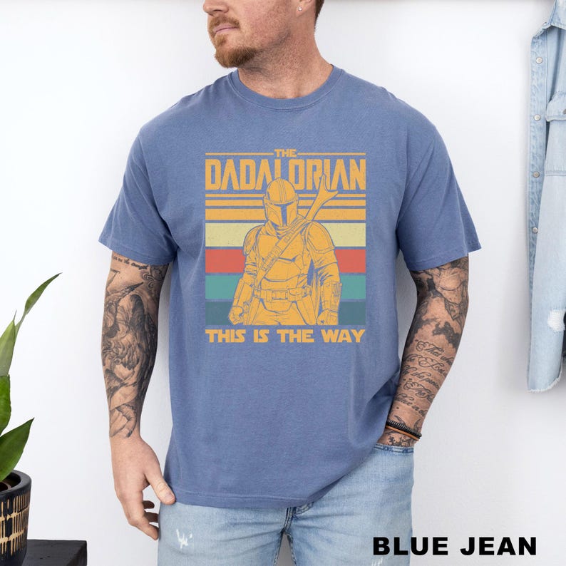 May include: Blue t-shirt featuring a graphic of a Mandalorian-style character with the text "THE DADALORIAN" and "THIS IS THE WAY" in a retro design. The shirt is a solid blue color.