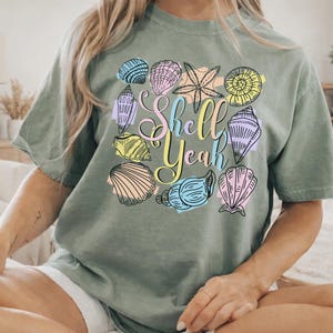 May include: A sage green t-shirt with a colorful graphic of seashells and the words "Shell Yeah". The seashells are in various colors, including blue, pink, yellow, and purple. The shirt has short sleeves and a relaxed fit.