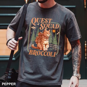 May include: Dark gray t-shirt featuring a cartoon cat with a backpack, surrounded by trees and the text "QUEST SQUAD BROCCOLI". The design includes the phrase "No One Wants It, But It's Good For You".