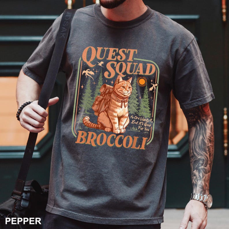 May include: Dark gray t-shirt featuring a cartoon cat with a backpack, surrounded by trees and the text "QUEST SQUAD BROCCOLI". The design includes the phrase "No One Wants It, But It's Good For You".