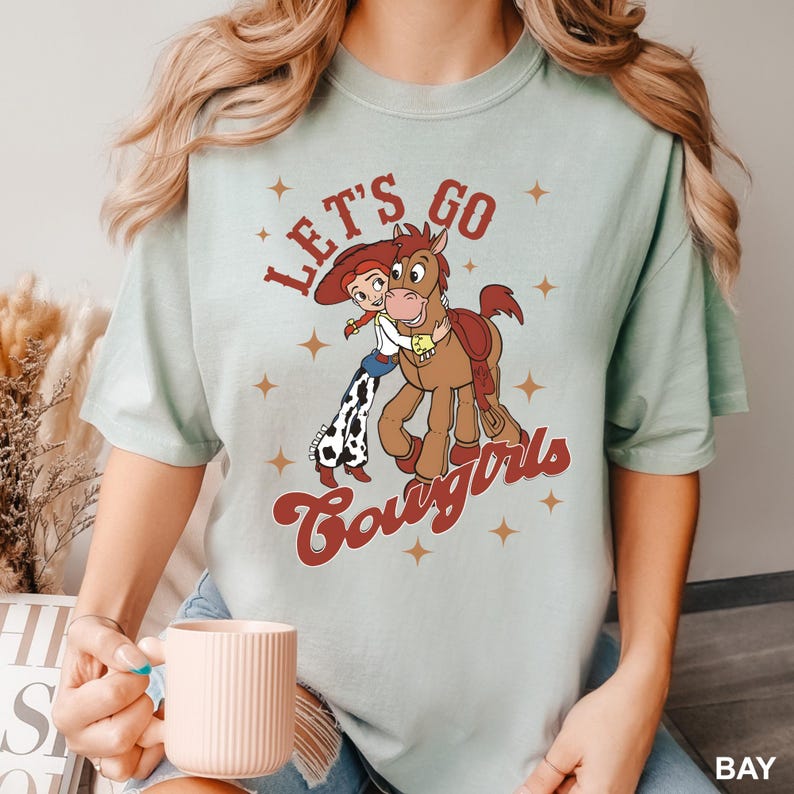 May include: A light blue t-shirt featuring a cartoon cowgirl and horse graphic. The text "Let's Go Cowgirls" is printed in red on the shirt.