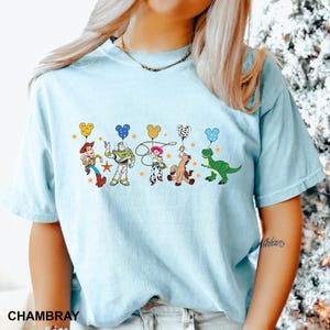 K&ouml;nnte beinhalten: Ein hellblaues T-Shirt mit einem Aufdruck von Toy Story-Charakteren, die Mickey-Maus-Ballons halten. Die Charaktere sind Woody, Buzz Lightyear, Jessie, Bullseye und Rex.
