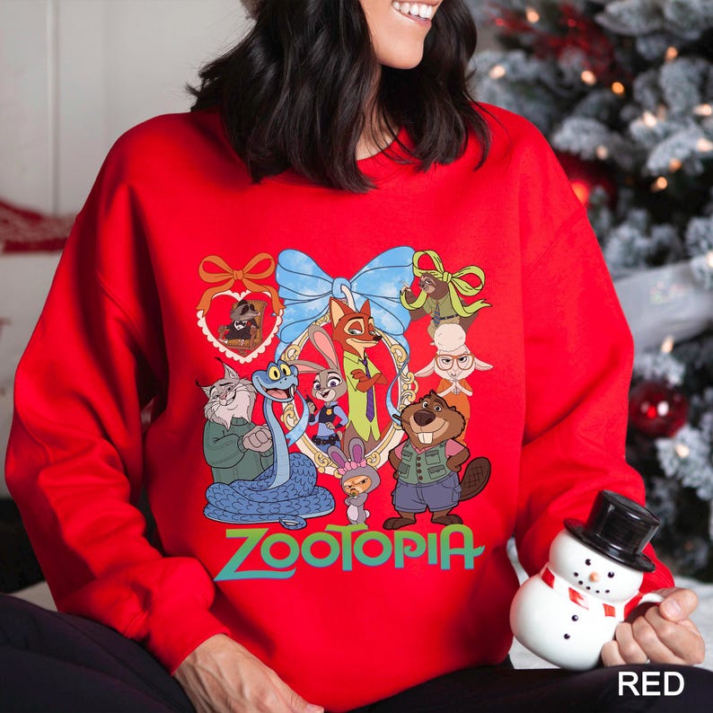 Op de afbeelding: Een rode sweatshirt met een kleurrijke afbeelding van personages uit de animatiefilm "Zootopia". Het ontwerp bevat een blauwe strik, ingelijste karakterportretten en het woord "Zootopia" in het groen. Een sneeuwpopfiguur staat op de voorgrond.