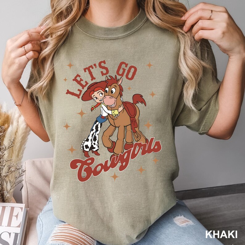 May include: A khaki green t-shirt featuring a vintage-style graphic of a cartoon cowgirl and horse. The graphic says "Let's Go Cowgirls".