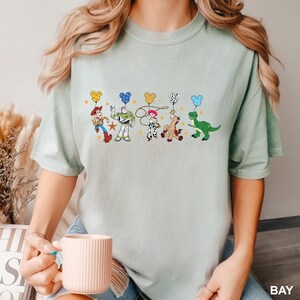 K&ouml;nnte beinhalten: Ein hellblaues T-Shirt mit einem Aufdruck von Toy Story-Charakteren, die Ballons in Form von Mickey-Maus-K&ouml;pfen halten. Die Charaktere sind Woody, Buzz Lightyear, Jessie, Bullseye und Rex.