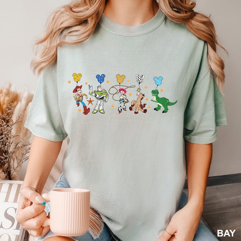 K&ouml;nnte beinhalten: Ein hellblaues T-Shirt mit einem Aufdruck von Toy Story-Charakteren, die Ballons in Form von Mickey-Maus-K&ouml;pfen halten. Die Charaktere sind Woody, Buzz Lightyear, Jessie, Bullseye und Rex.